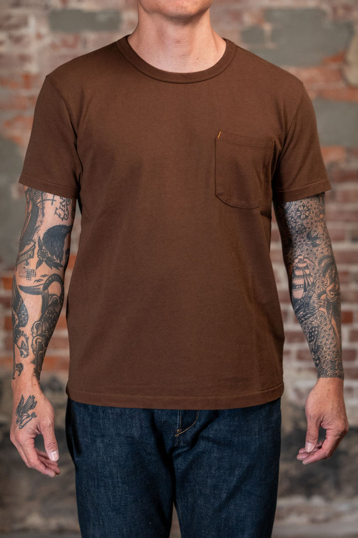 A man wearing the 
Freenote Cloth 13oz Pocket T-Shirt - Chocolate, a  brown short-sleeve crewneck pocket t-shirt and dark blue jeans, standing in front of a rustic brick wall background.