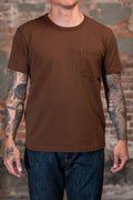 A man wearing the 
Freenote Cloth 13oz Pocket T-Shirt - Chocolate, a  brown short-sleeve crewneck pocket t-shirt and dark blue jeans, standing in front of a rustic brick wall background.
