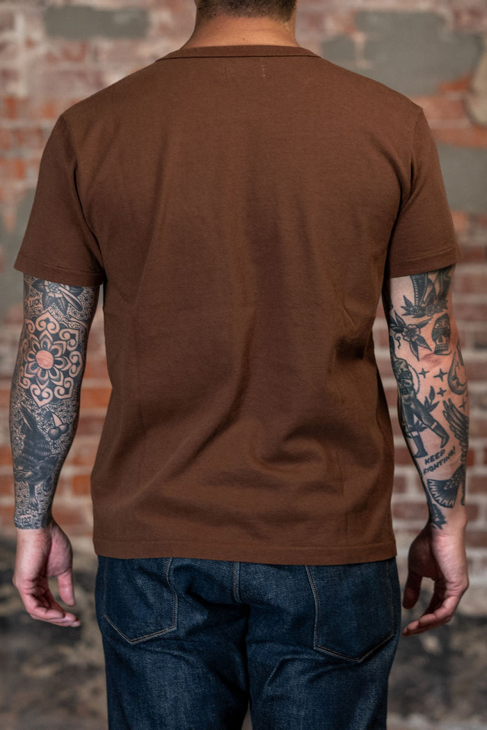 A man viewed from behind wearing the 
Freenote Cloth 13oz Pocket T-Shirt - Chocolate, a solid brown short-sleeve t-shirt and blue jeans, standing against a rustic brick wall background.