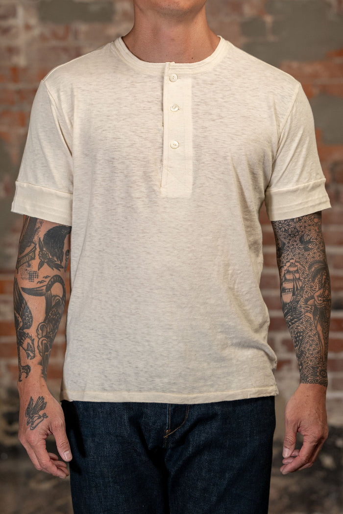 A man wearing the Freenote Cloth Fairmont Henley S/S - Natural - backlight beige heathered short-sleeve henley t-shirt with rolled cuffs and three buttons, paired with dark blue jeans, standing against a rustic brick wall.