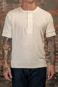 A man wearing the Freenote Cloth Fairmont Henley S/S - Natural - backlight beige heathered short-sleeve henley t-shirt with rolled cuffs and three buttons, paired with dark blue jeans, standing against a rustic brick wall.
