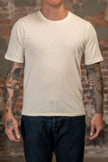 A man wearing the 
Freenote Cloth Fairmont S/S - Natural a light beige crew-neck t-shirt and dark blue jeans, standing in front of a rustic brick wall background.
