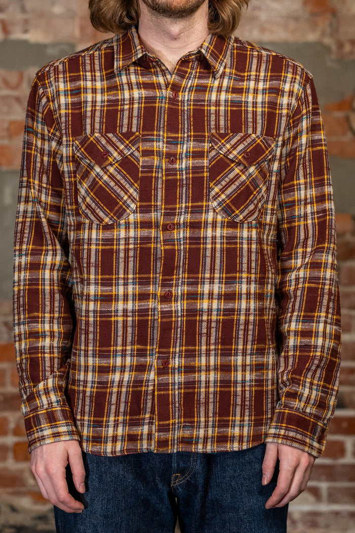 A man wearing the 
UES 502655 Slubby Flannel Shirt - Brown, a brown, yellow, and white plaid button-up flannel shirt, paired with blue jeans, standing in front of a brick wall.