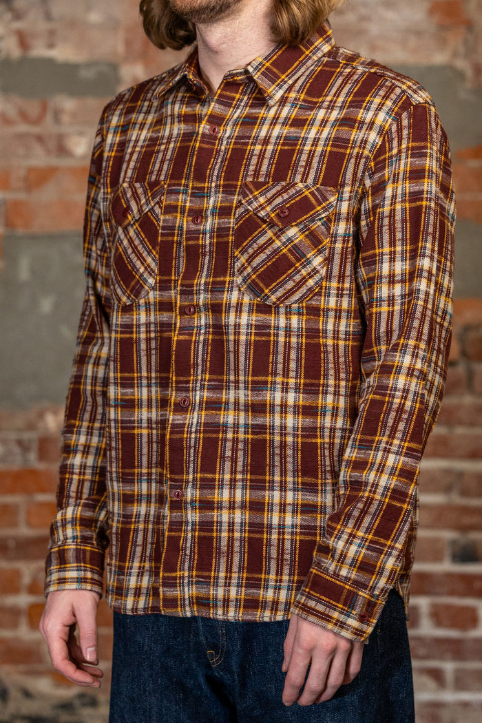 A man wearing the 
UES 502655 Slubby Flannel Shirt - Brown, a  brown, yellow, and white plaid long-sleeve button-up shirt, paired with dark blue jeans, standing in front of a rustic brick wall.