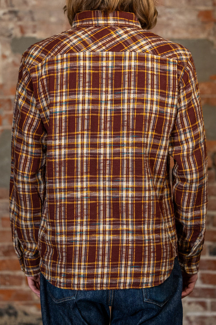 A person viewed from the back wearing the 
UES 502655 Slubby Flannel Shirt - Brown, a long-sleeve plaid button-up shirt in brown, mustard yellow, and white, paired with blue jeans, set against a rustic brick wall background.