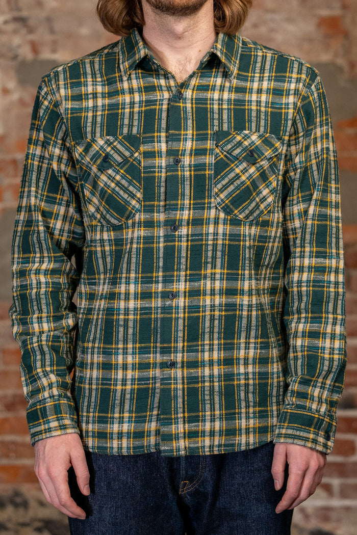A man wearing 
UES 502655 Slubby Flannel Shirt - Green, a green and yellow plaid long-sleeve button-up shirt with two chest pockets, paired with dark blue jeans, standing against a rustic brick wall.