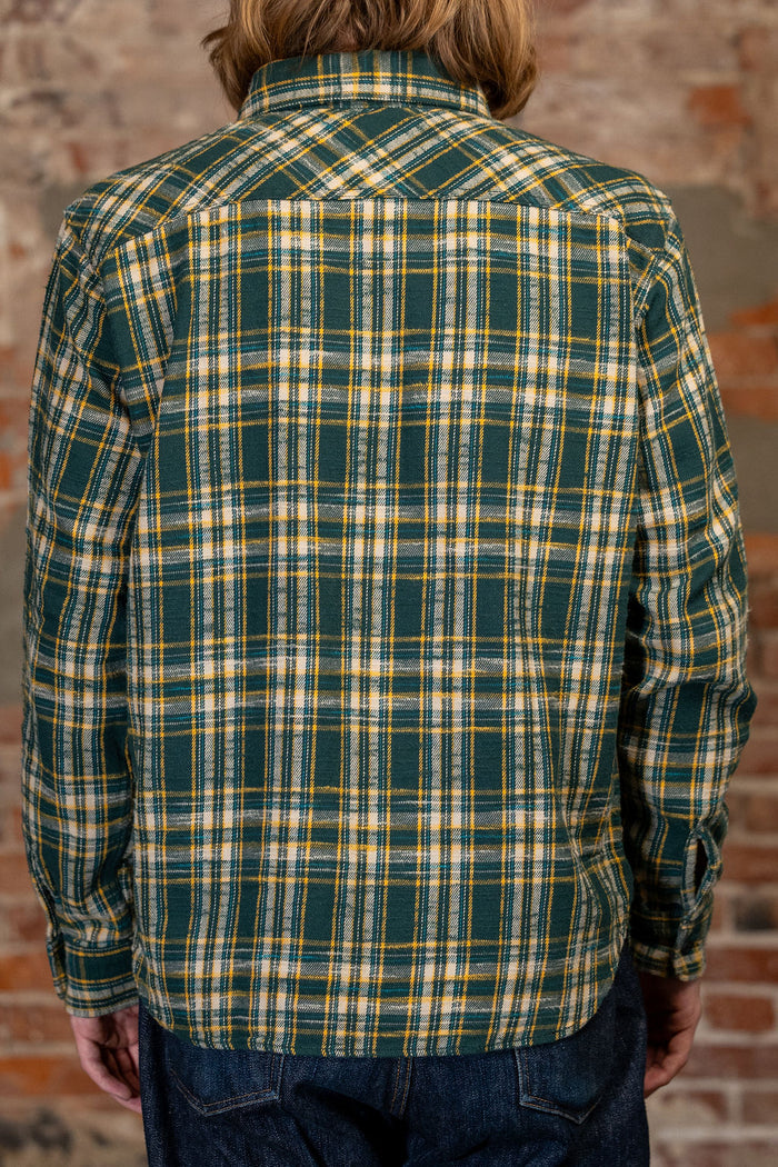 A person viewed from the back wearing 
UES 502655 Slubby Flannel Shirt - Navy  a green plaid flannel shirt with yellow and white stripes, paired with dark blue jeans, set against a rustic brick wall background.