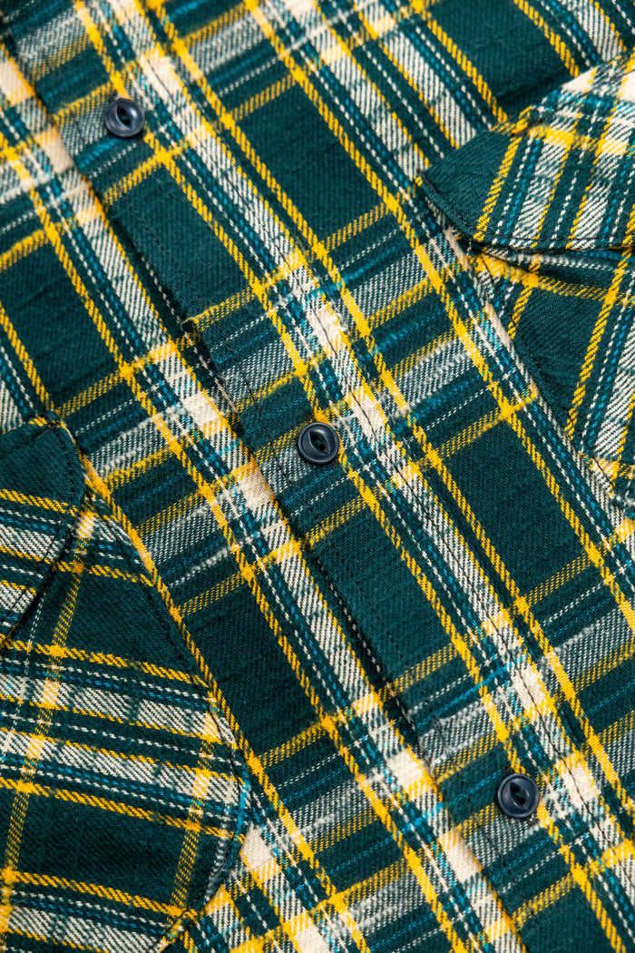 Close-up of a green plaid flannel shirt with yellow and white stripes, featuring dark buttons and a visible collar. 
UES 502655 Slubby Flannel Shirt - Green
