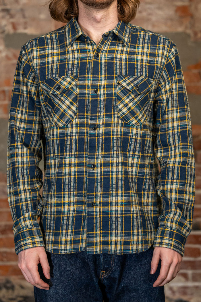 A man wearing 
UES 502655 Slubby Flannel Shirt - Navy a long-sleeve button-up flannel shirt with a navy blue, yellow, and white plaid pattern, paired with dark blue jeans, standing against a rustic brick wall.