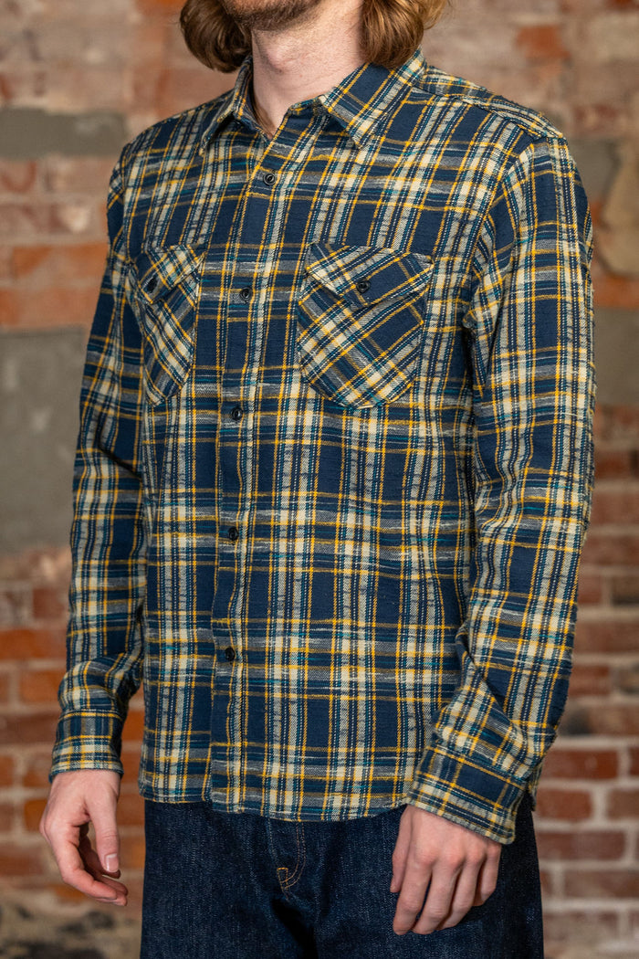 A man wearing 
UES 502655 Slubby Flannel Shirt - Navy a long-sleeve button-up plaid shirt in navy blue, yellow, and white, paired with dark blue jeans, standing against a rustic brick wall.