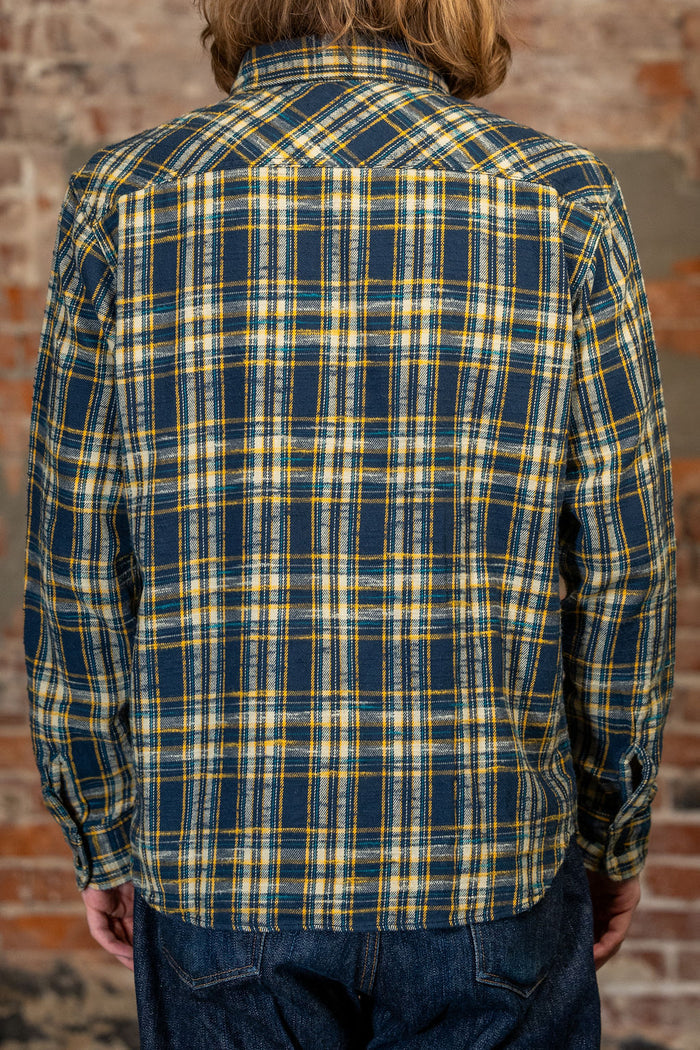 A person viewed from the back wearing the 
UES 502655 Slubby Flannel Shirt - Navy, a long-sleeve plaid button-up shirt in dark blue with yellow and white stripes, paired with dark blue jeans, set against a rustic brick wall background.