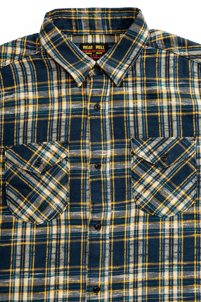 UES 502655 Slubby Flannel Shirt - Navy A close-up of a long-sleeve plaid button-up shirt by Wear Well, featuring a navy blue, yellow, and white tartan pattern with two chest pockets.