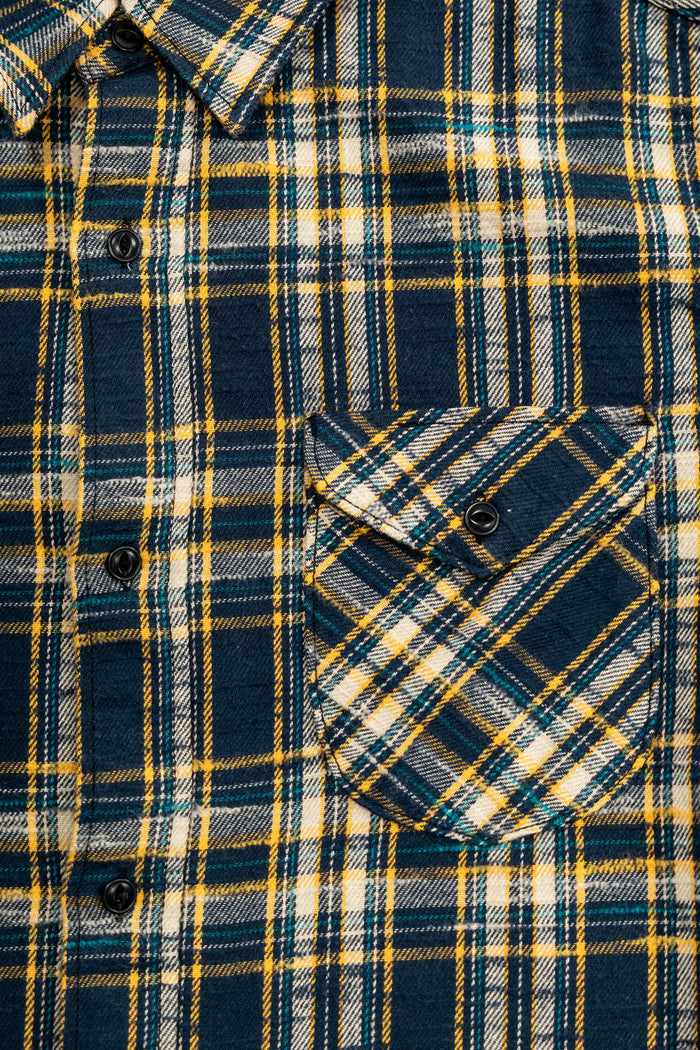 UES 502655 Slubby Flannel Shirt - Navy A close-up of a plaid button-up shirt with a chest pocket, featuring navy blue, yellow, and white stripes.