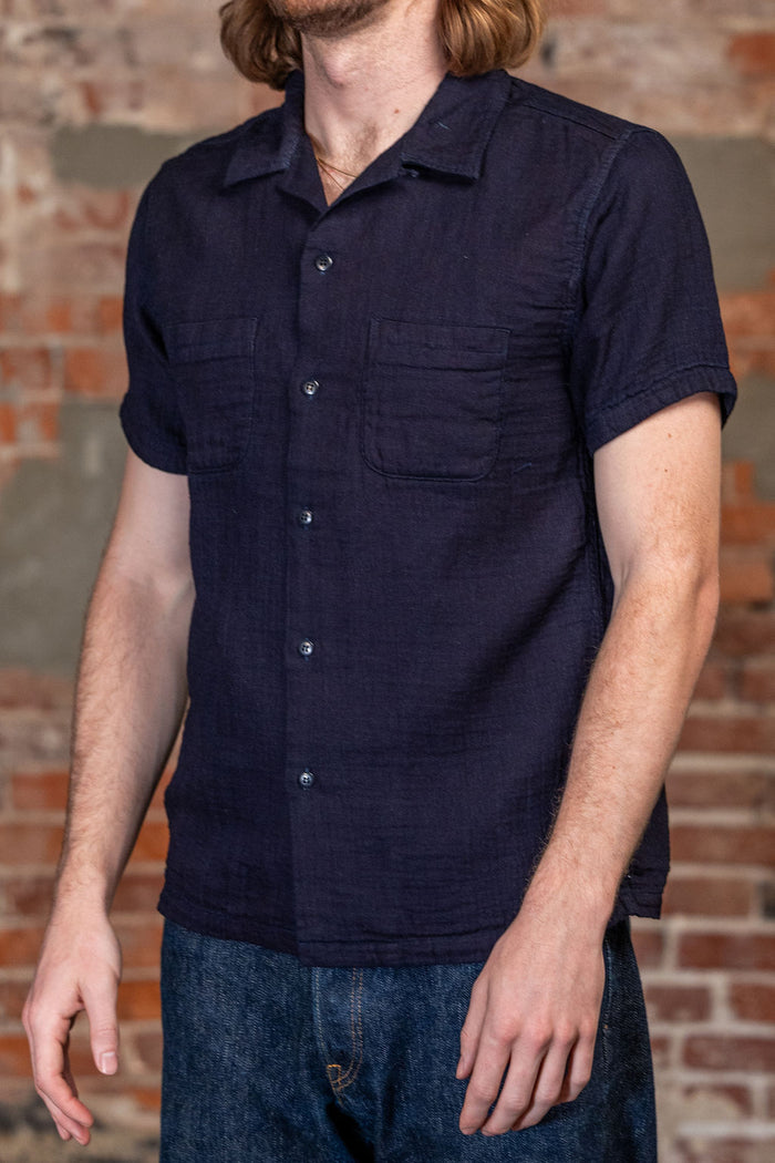 A man wearing the 
UES Herringbone Double Gauze Short Sleeve Shirt - Indigo  dark navy blue short-sleeve button-up shirt with two chest pockets, paired with blue jeans, standing in front of a rustic brick wall.