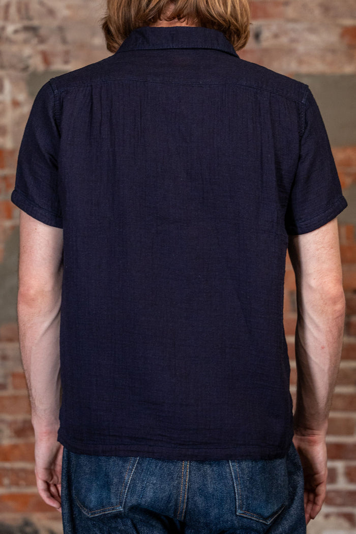 A man viewed from behind wearing 
UES Herringbone Double Gauze Short Sleeve Shirt - Indigo  a dark navy blue short-sleeve linen shirt and blue jeans, standing in front of a rustic brick wall.