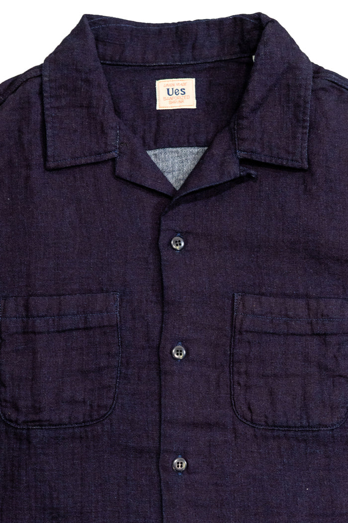 UES Herringbone Double Gauze Short Sleeve Shirt - Indigo: A close-up of a dark indigo blue cotton shirt jacket with a collar, button front, and two chest pockets. A brand label reading 'Ues' is visible inside the collar.