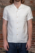 A man wearing the 
UES Herringbone Double Gauze Short Sleeve Shirt - White, a white short-sleeve linen camp collar shirt with button-down front and two chest pockets, paired with dark blue jeans, standing in front of a rustic brick wall.