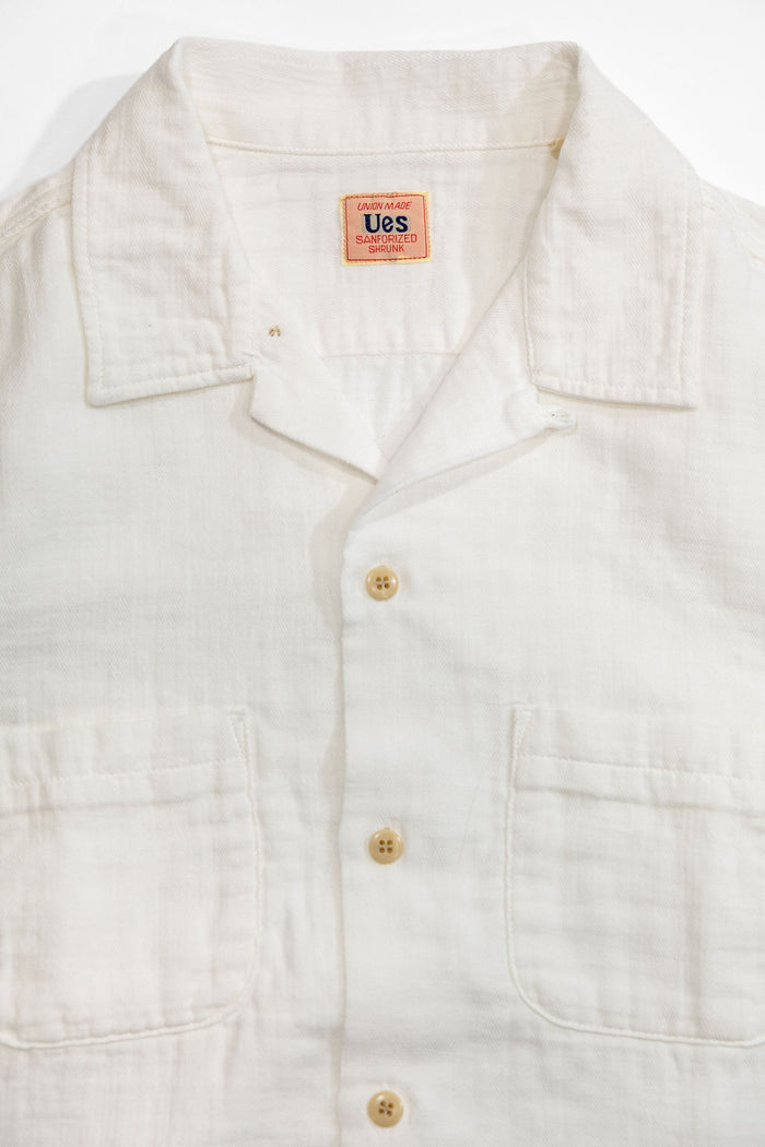 UES Herringbone Double Gauze Short Sleeve Shirt - White: A close-up of a white linen short-sleeve button-up shirt with a collar, two front chest pockets, and light tan buttons. The inner neck label shows the brand 'Ues'.